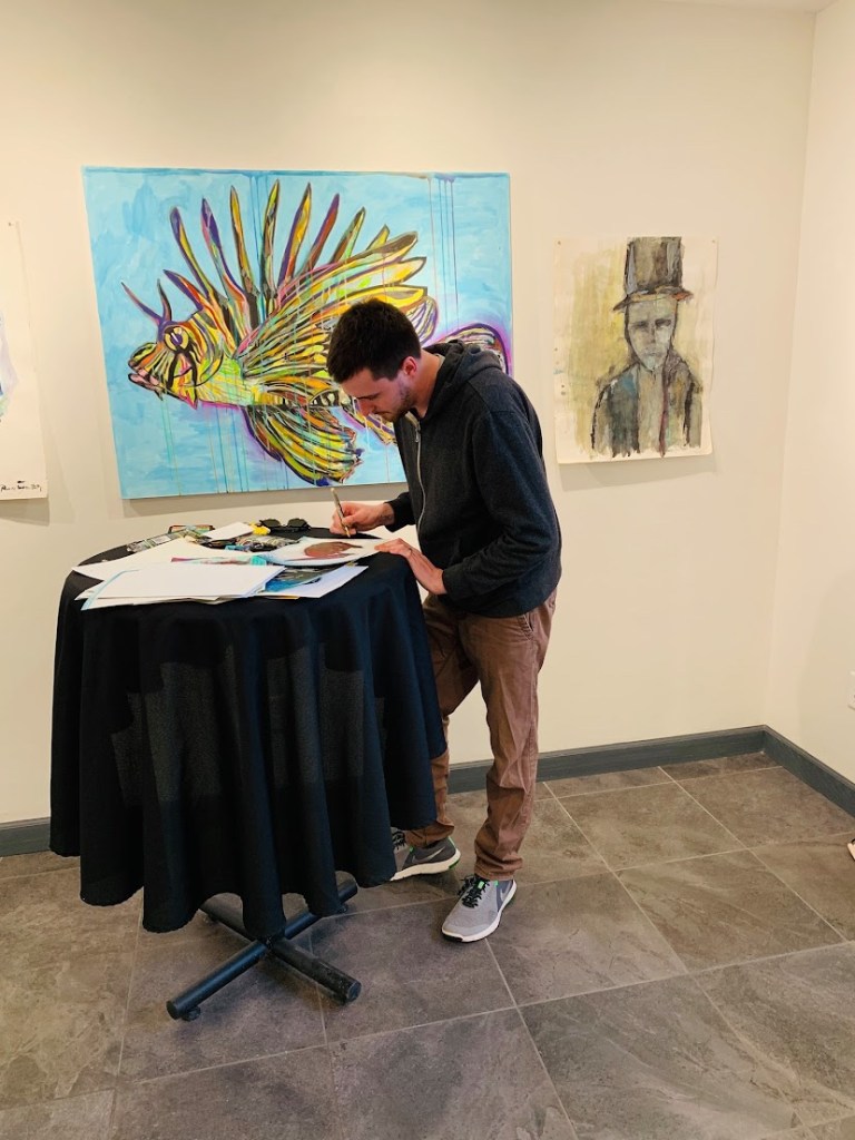Artist Russell Frantom (south bend Indiana / Chicago) stands near his lionfish painting while he signs lithographs for his art show. 2019